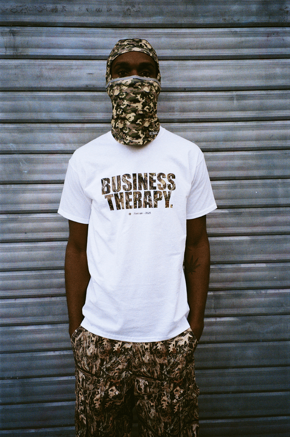 Homme cagoulé devant un rideau de fer, portant le Business Therapy Camo T-shirt, plan resserré sur le torse.