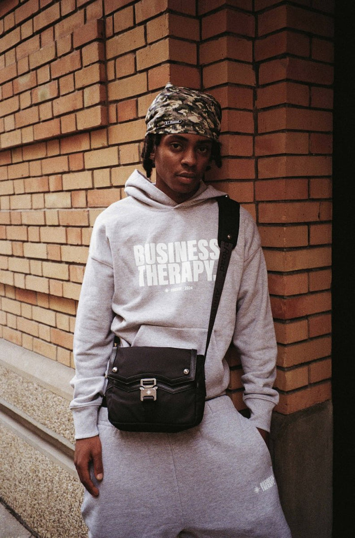 "Homme en jogging gris Business Therapy avec Memorial Bag, au coin d’une rue devant mur de briques