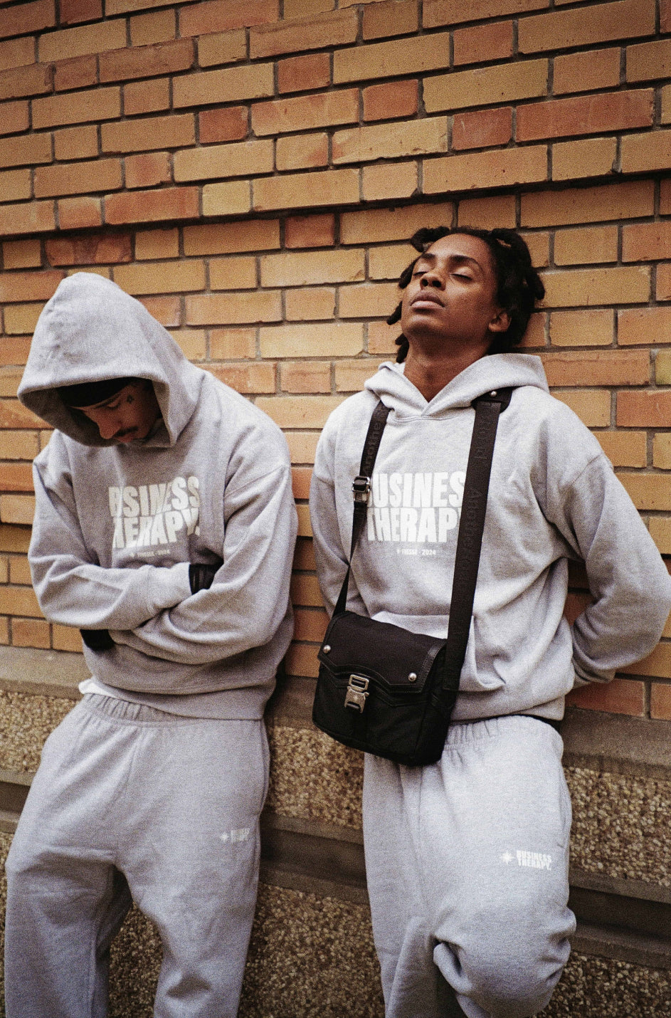 Deux hommes adossés à un mur de briques, en jogging gris Business Therapy, avec capuche et Memorial Bag