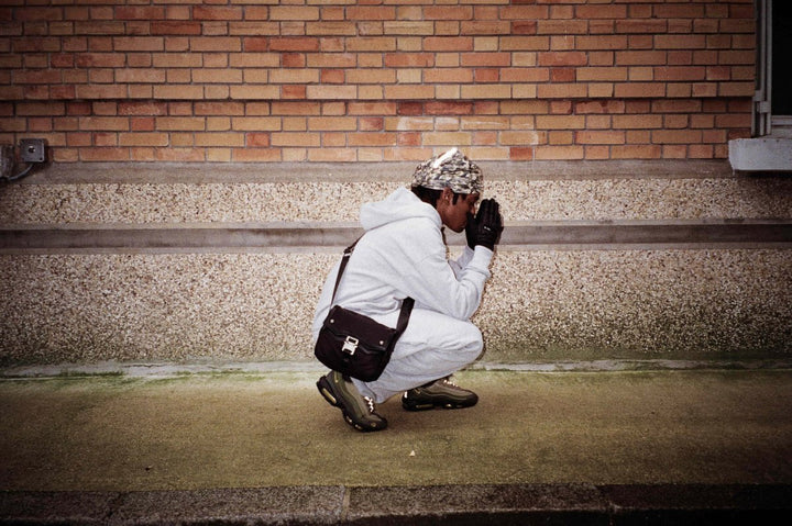 Homme accroupi en jogging gris Business Therapy avec Memorial Bag, mains jointes comme en prière.