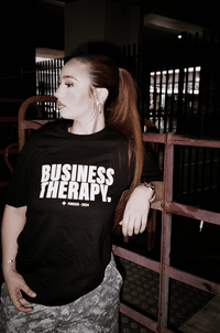 Femme dans un décor urbain portant un t-shirt noir Business Therapy, regard tourné vers la droite.