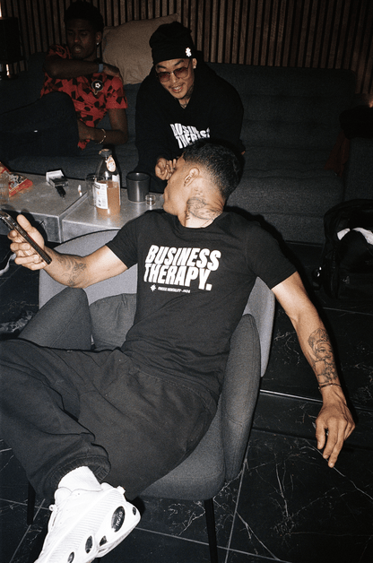 Homme tatoué allongé dans un canapé, portant un t-shirt Business Therapy et des sneakers blanches.