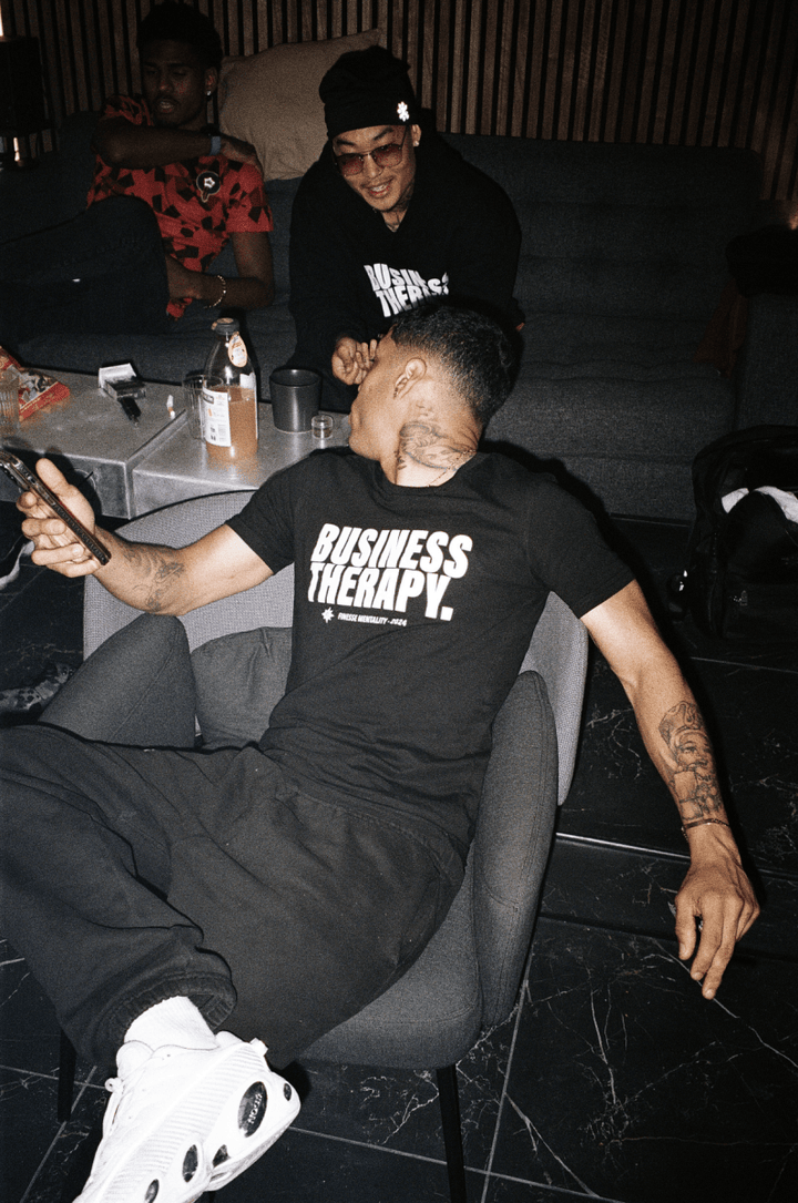 Homme tatoué allongé dans un canapé, portant un t-shirt Business Therapy et des sneakers blanches.