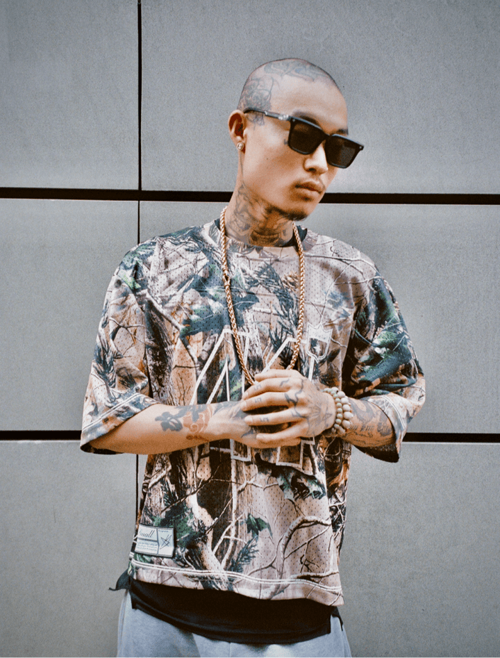 Homme tatoué en Jersey 44 camo, lunettes Sunglasses Series 01, mains jointes.