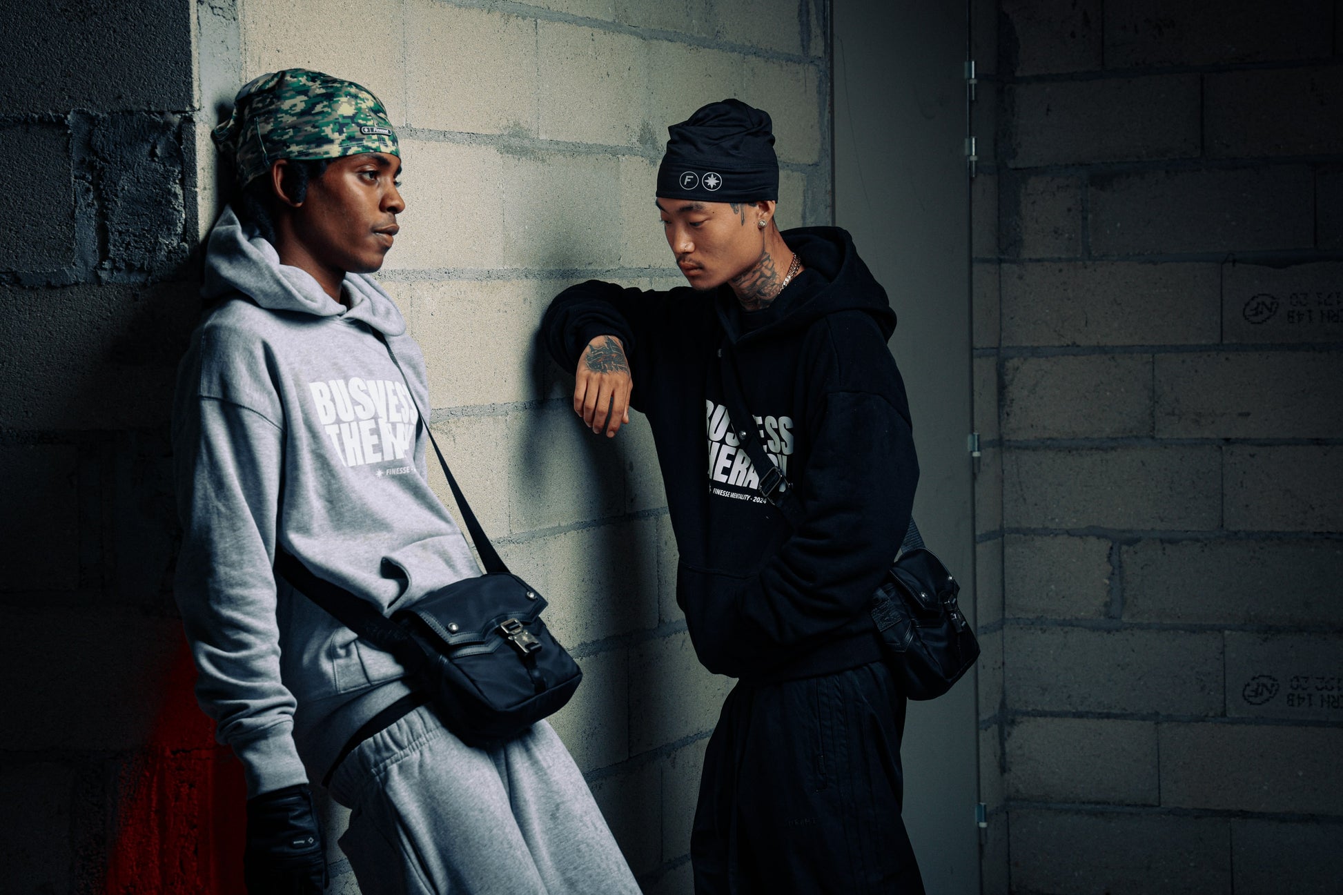 Deux hommes adossés à un mur en jogging noir Business Therapy, tous deux avec Memorial Bag. (variante)