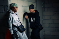 Deux hommes adossés à un mur en jogging noir Business Therapy, tous deux avec Memorial Bag. (variante)