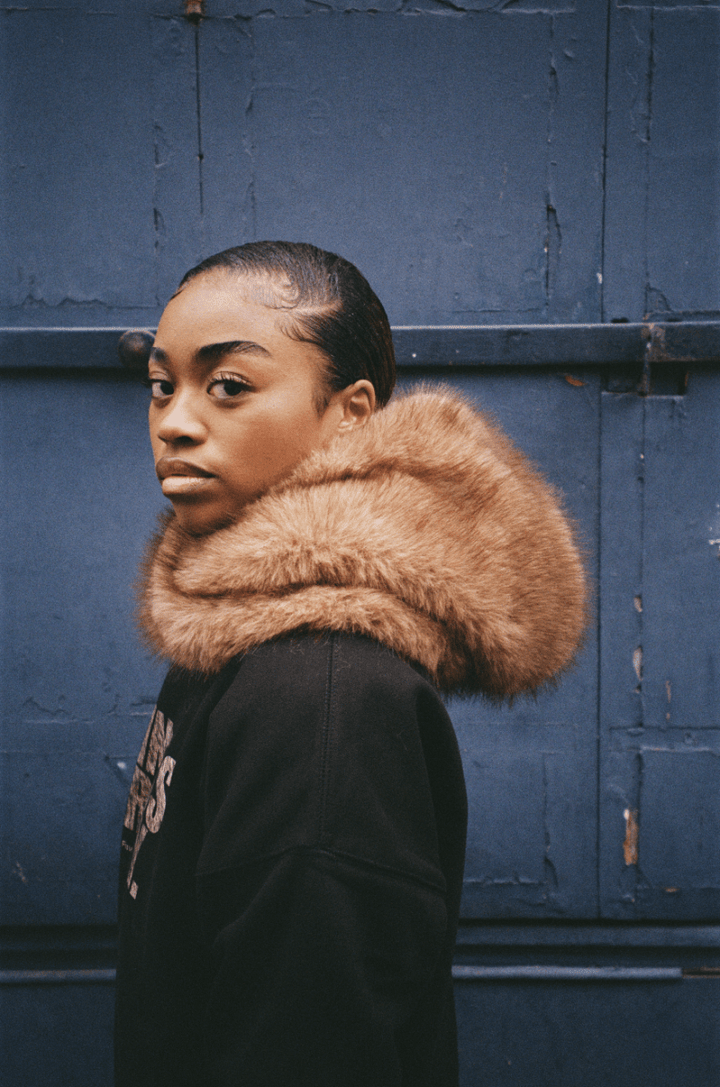 Personne de profil devant une porte bleue, capuche FUR HOOD abaissée.