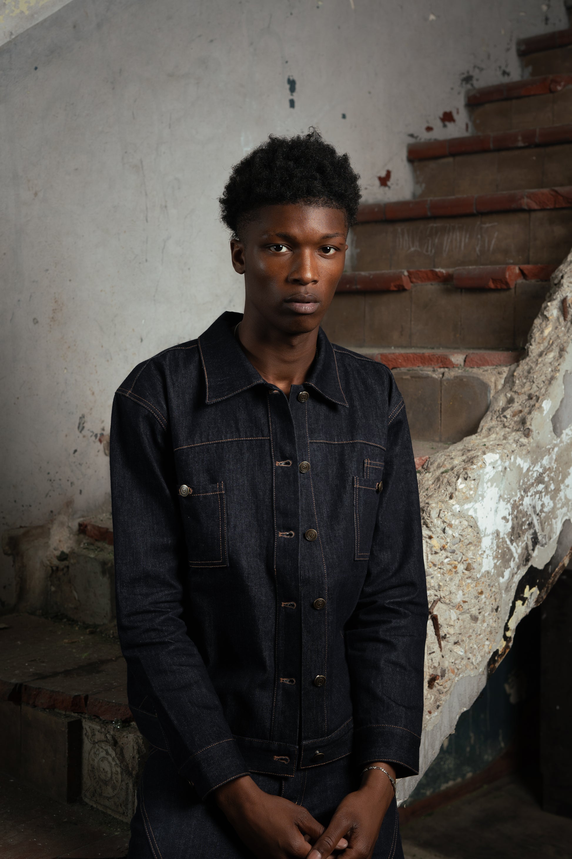 Homme posant dans un bâtiment abandonné, portant la Finesse Denim Jacket ouverte près d’un escalier.