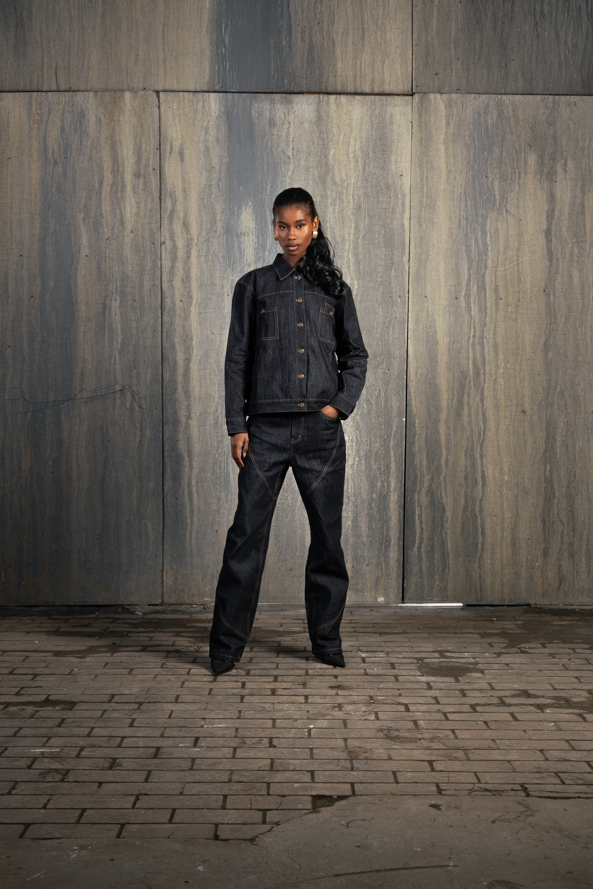 Femme de face en studio décor industriel, main dans la poche, portant le Finesse Denim Set ADV avec allure affirmée.