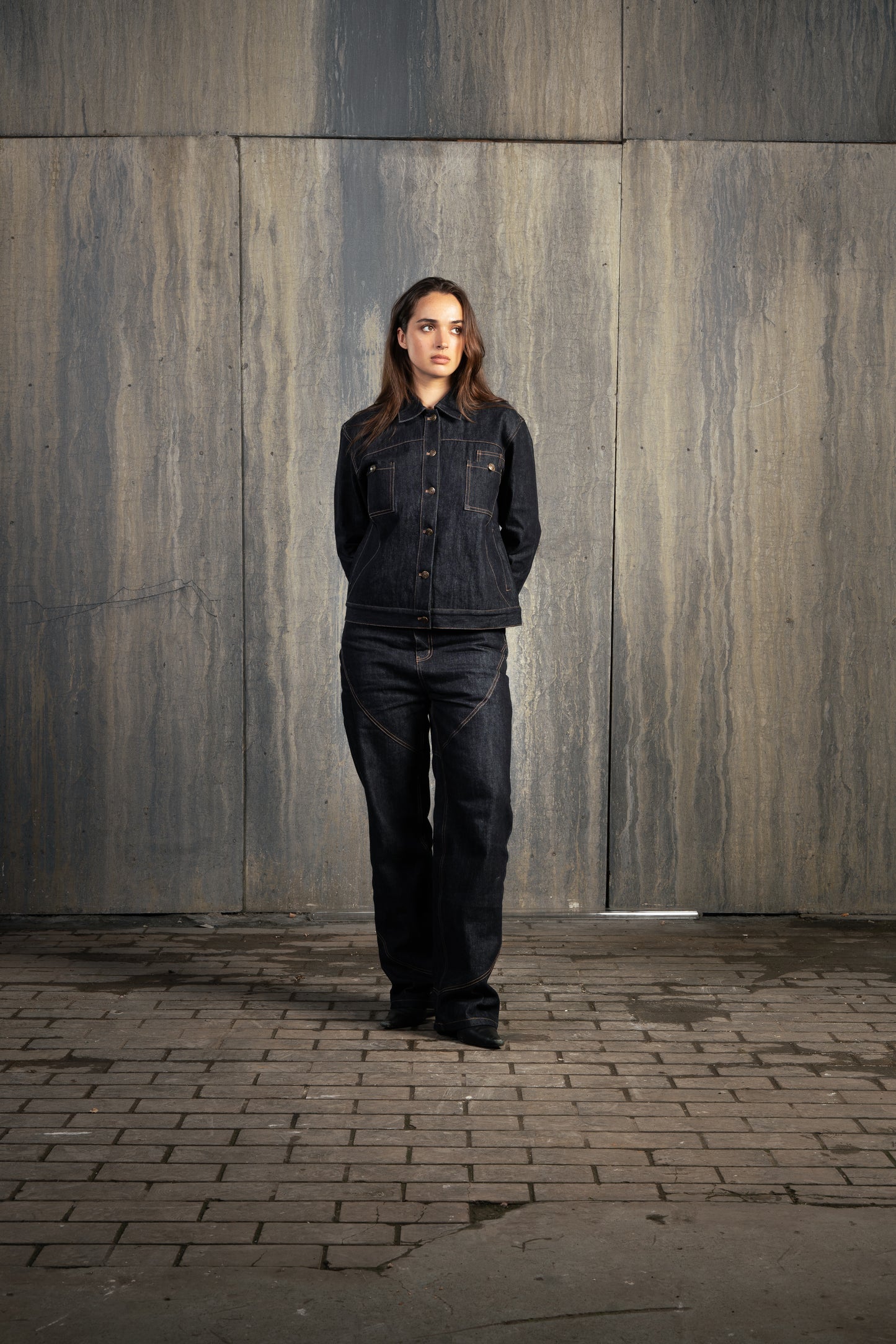 Femme en studio décor industriel, regard malicieux vers la gauche, mains derrière le dos, portant l’ensemble Finesse Denim Set ADV.