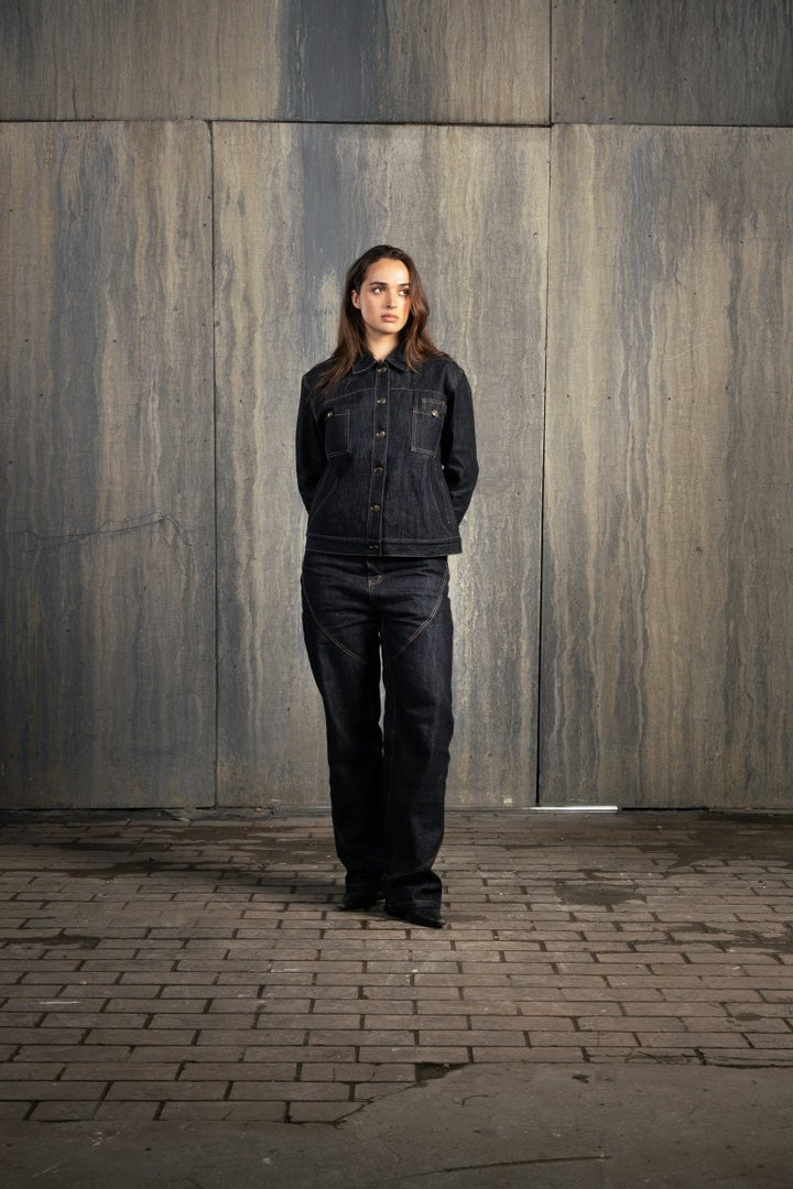 Femme en studio décor industriel, regard malicieux vers la gauche, mains derrière le dos, portant l’ensemble Finesse Denim Set ADV.