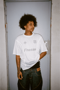 Homme tatoué devant une porte portant un Finest Denim Short et un Finesse Jersey Shirt blanc.