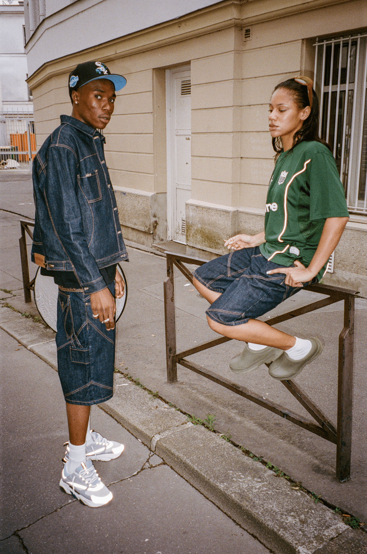Femme assise sur une barrière en Finest Denim Short et Jersey vert, avec un homme en Denim Short, veste et casquette.