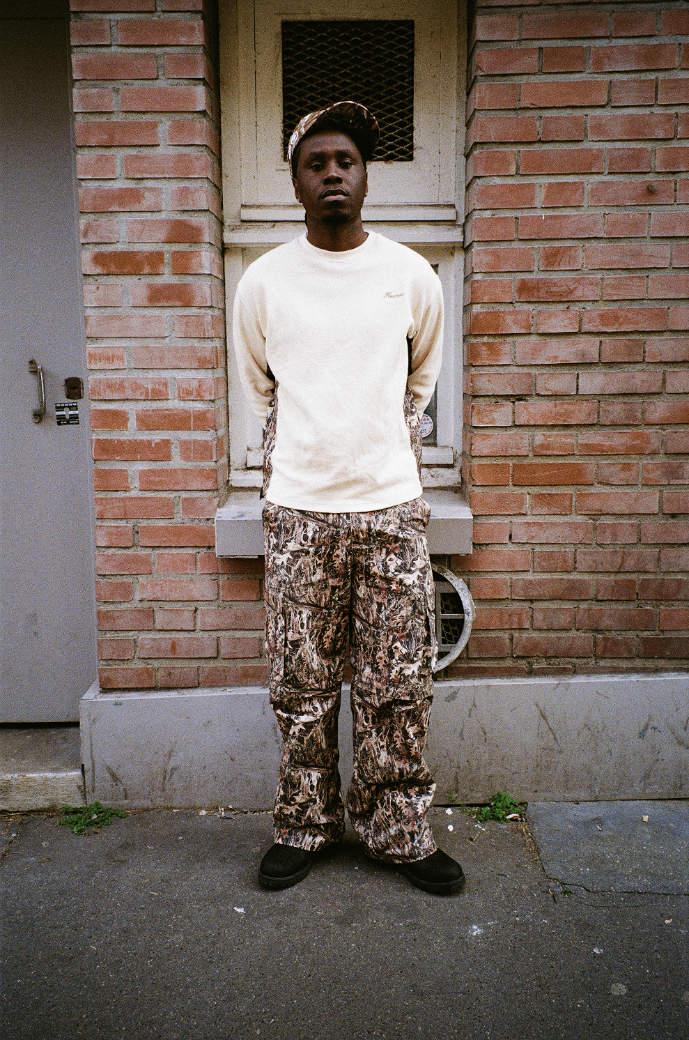 Rappeur KAINZ devant mur de briques, portant Finesse Military Top-Camo, 2-in-1 Cargo Pants-Camo et "SIGN" Cap-CAMO.