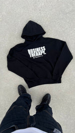 Hoodie Business Therapy Black de Finesse en packshot à plat, avec les pieds du photographe visibles.