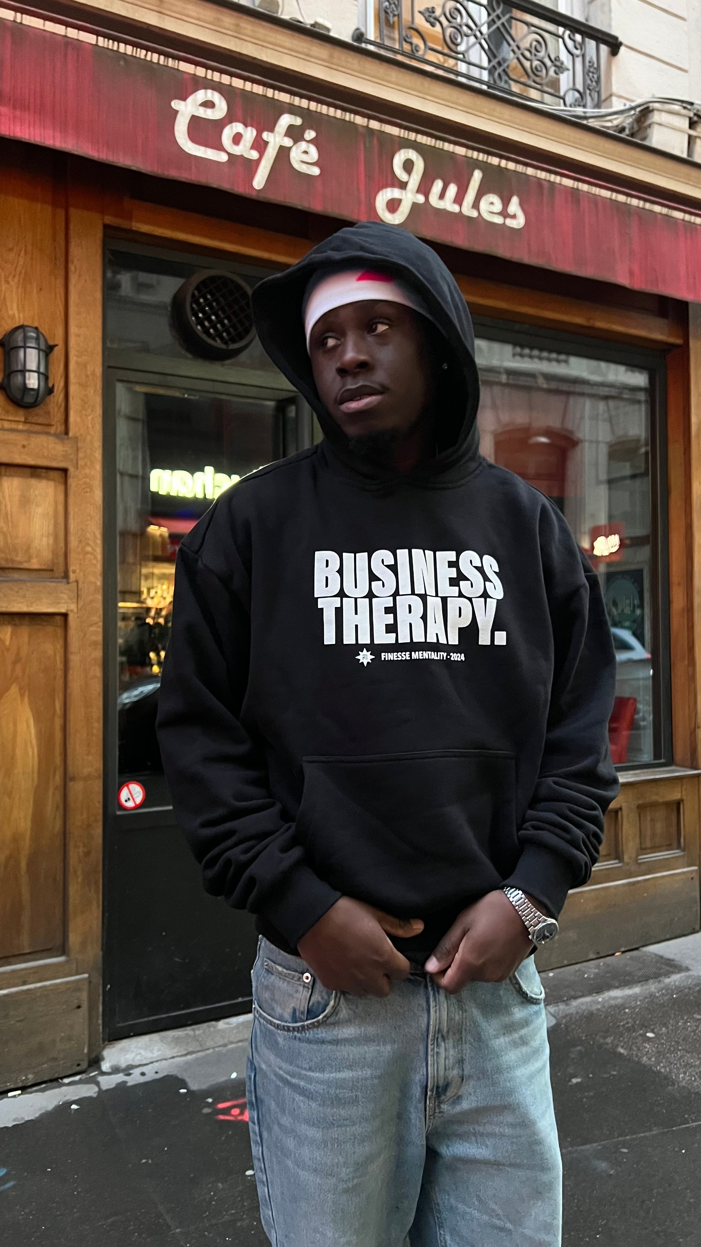 Rappeur devant la façade Café Jules portant un hoodie Business Therapy Finesse et une montre.