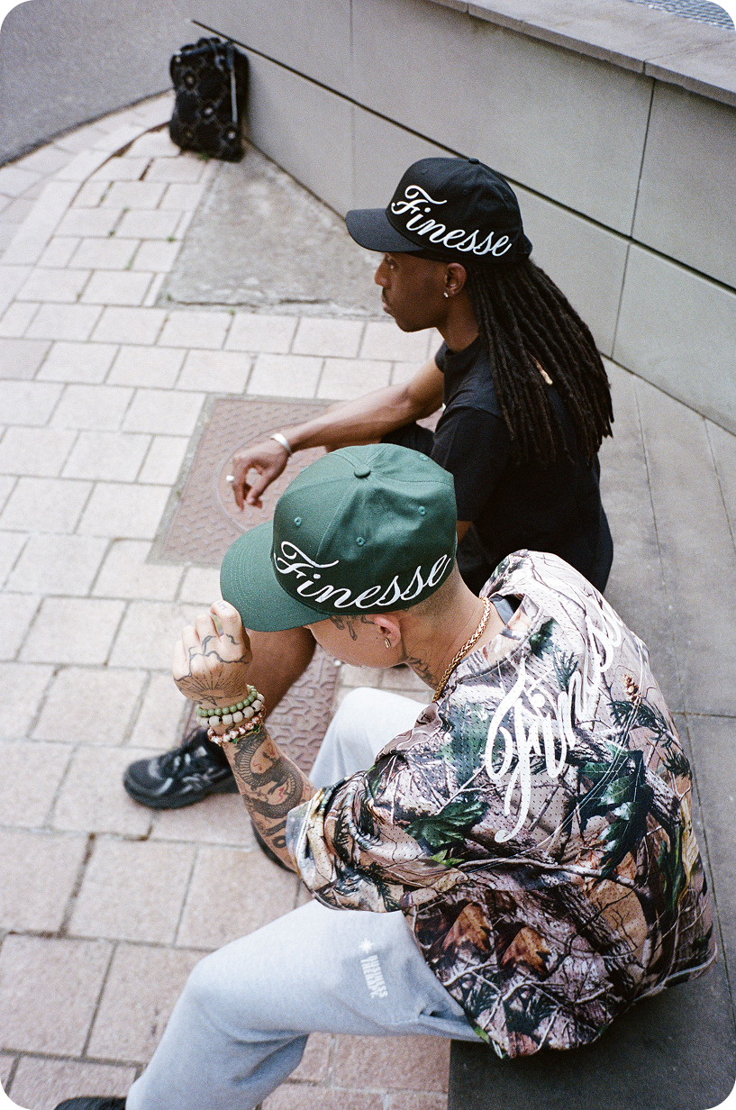 Deux hommes assis dans la rue, l’un en Jersey 44 camo, l’autre en noir, casquettes Sign Cap verte et noire