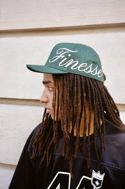 Homme portant une Finesse Sign Cap verte, regard à droite devant un mur.