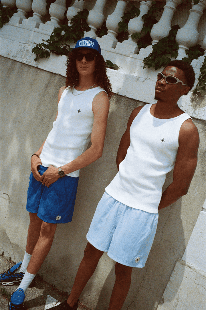 Deux hommes adossés à un mur, en Tank Top, Swim Shorts et casquettes Business Therapy.